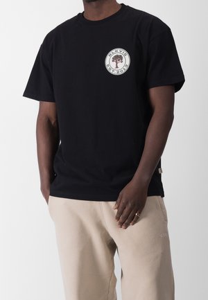 Czarna bawełniana koszulka z krótkim rękawem z okrągłym logo z motywem drzewa oraz napisem "BLKVIS EST. 2013." Luźny krój, noszona z beżowymi spodniami.