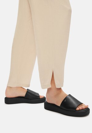 Sabe de piele negre, cu design minimalist, formă rectangulară și talpă ridicată. Complementate de pantaloni bej, cu croială lejeră.