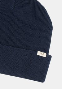 Berretto lavorato a maglia blu navy realizzato in materiale morbido e testurizzato. Presenta un risvolto e una piccola etichetta in tessuto con un logo sul lato.