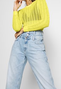 Ljusgul ribbad långärmad topp med dekorationer, ihop med högmidjade ljusblå jeans i denim med knappstängning och avslappnad passform.