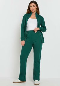 Groene sportjack met rits, gecombineerd met bijpassende broek. Beide gemaakt van zachte stof, met een ontspannen pasvorm en elastiek in de taille. Witte top en sneakers.