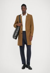 Palton din lână maro cu rever tăiat, nasturi negri, cămașă albă, pantaloni bleumarin și o geantă din piele neagră. Textură netedă, design croit.