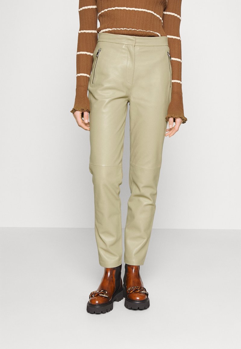 Personne portant un pantalon en cuir beige avec des poches zippées à l'avant, un haut à manches longues rayé marron et des bottes épaisses marron avec un détail chaîne.