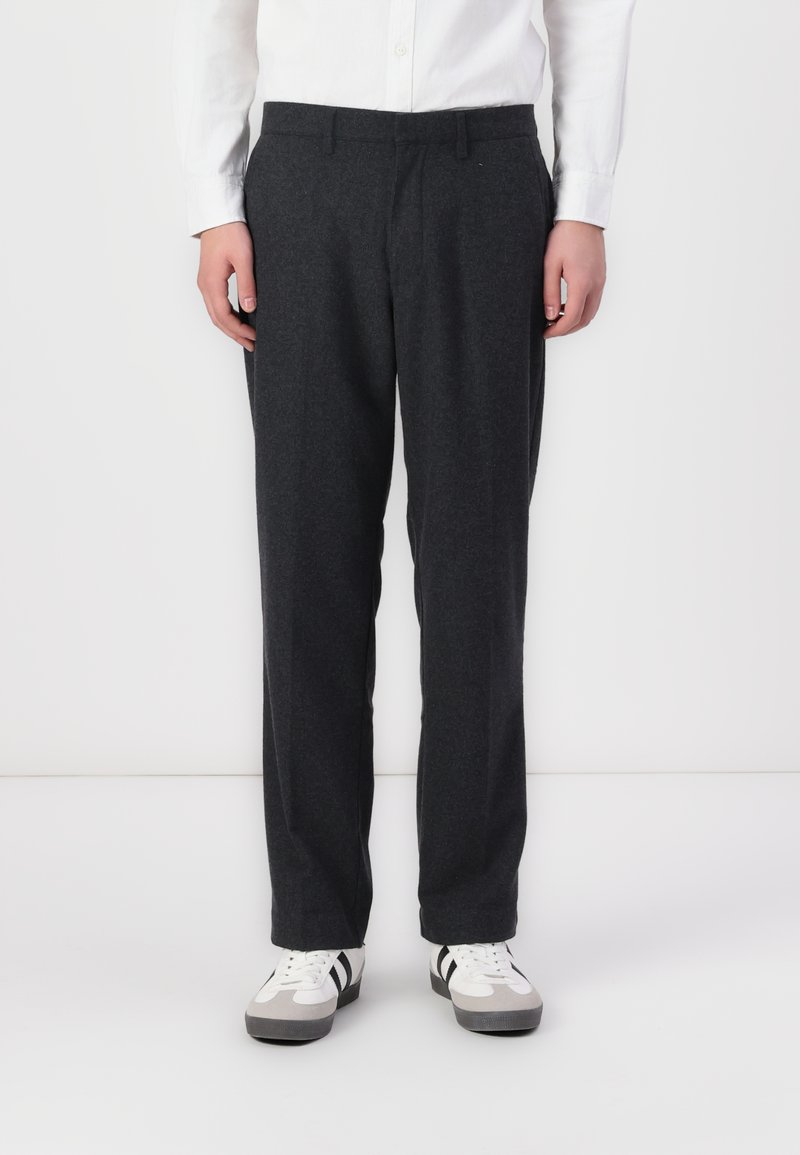 Pantalons en laine grise à coupe droite, avec un devant plat et des poches latérales. Portés avec des baskets blanches et grises.