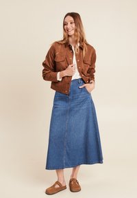 Veste cropped en daim marron avec deux poches poitrine, portée sur une chemise blanche et une longue jupe en jean bleue, associée à des chaussures slip-on marron.