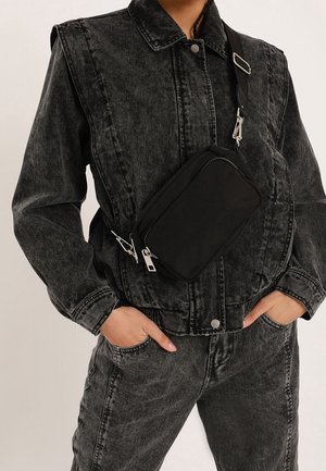 Persona que lleva puesta una chaqueta y pantalones vaqueros negros con lavado ácido, con las manos en los bolsillos, llevando un bolso cruzado negro con cremallera y hebilla plateadas.