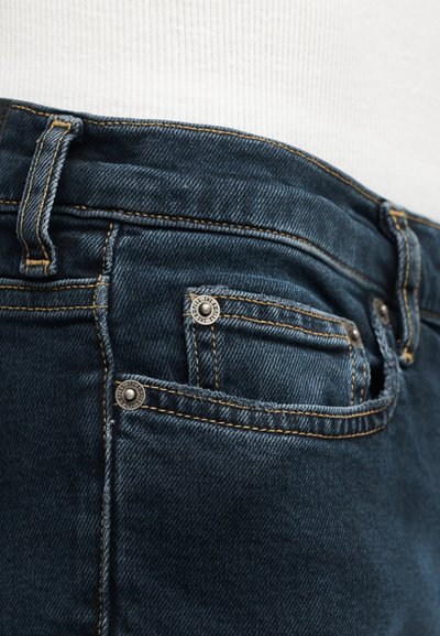 Κοντινή λήψη στην περιοχή της μπροστινής τσέπης σκούρων μπλε τζιν, με εμφανή ραφές και μεταλλικά καρφιά, πάνω σε λευκό ύφασμα φόντο.