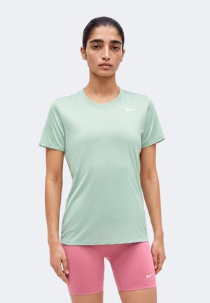 Femme debout, portant un t-shirt Nike vert clair à manches courtes et un short de cycliste Nike rose sur un fond clair uni.