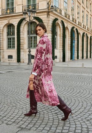 Manteau long à motif floral en rose et crème avec une fente latérale, associé à des bottes à talons hautes jusqu'aux genoux de couleur bordeaux foncé. Texture lisse, coupe décontractée.