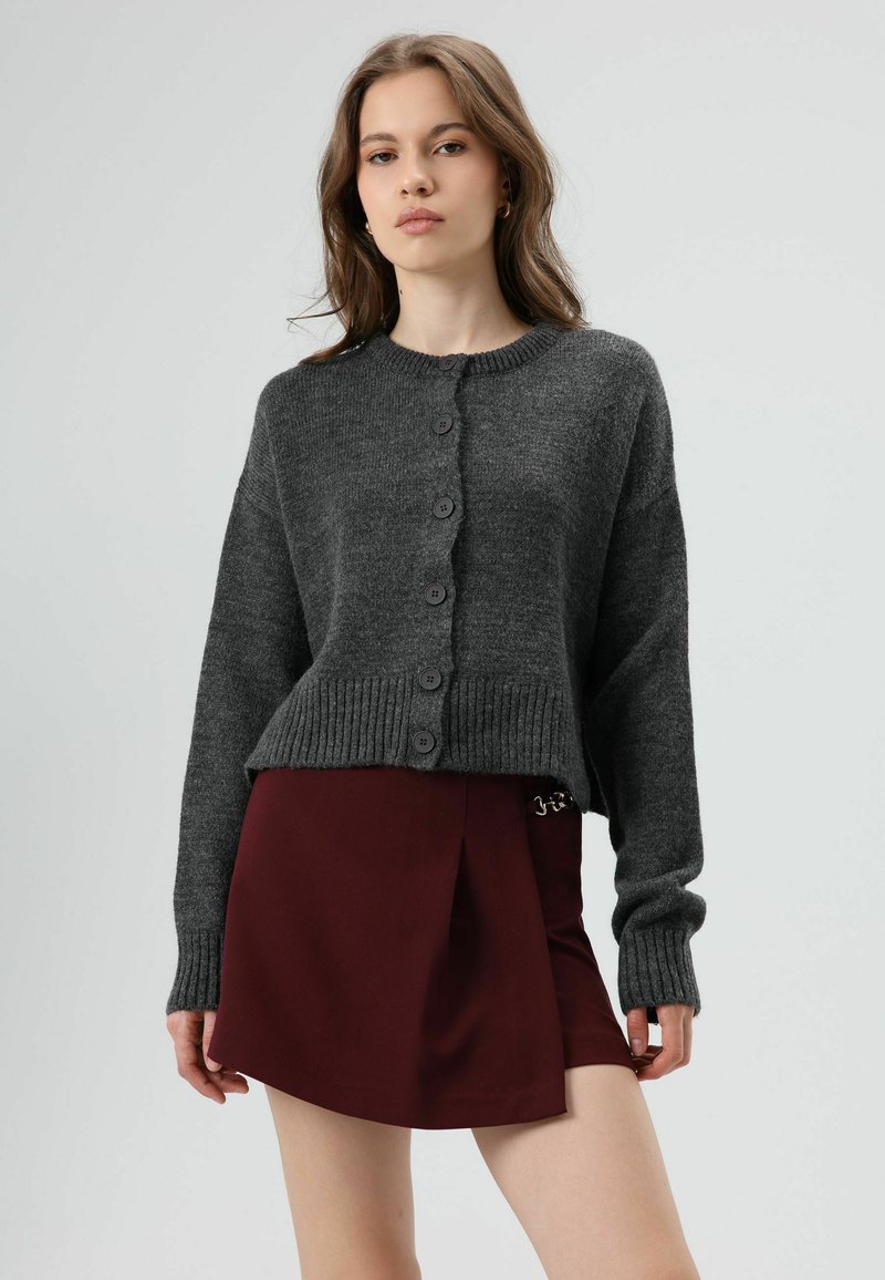 Cardigan en maille gris foncé avec un col rond, des poignets côtelés et une fermeture à boutons sur le devant, associé à une jupe bordeaux.