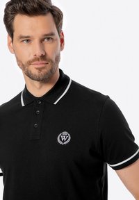 Černé polo tričko s bílými akcenty, s límečkem a třemi knoflíky. Obsahuje malé vyšívané logo na hrudi.