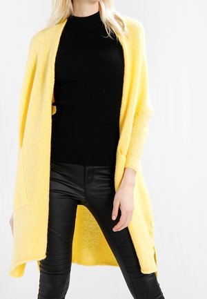 Kvinde iført en lang gul åben cardigan over en sort ribbet sweater og sorte læderbukser, stående mod en hvid baggrund.