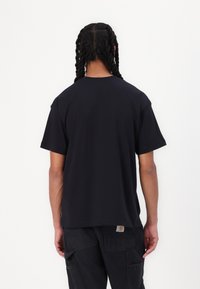 Black cotton t-shirt with short sleeves and a relaxed fit, featuring a visible logo tag on the lower side. Model is facing away from the camera.