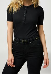 Musta ribbed lyhythihainen paita, jossa on nappilista, yhdistettynä mustiin korkea vyötäröisiin farkkuihin ja mustaan vyöhön, jossa on kultainen solki.