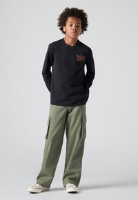 Μαύρο μακρυμάνικο πουκάμισο με λεπτομέρεια λογότυπου, συνδυασμένο με χαλαρά ελαιόχρωμα cargo παντελόνια με πλάγιες τσέπες και κρεμ sneakers.