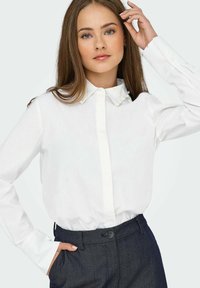 Chemise blanche à boutons avec des accents en perles au col, associée à un pantalon foncé ajusté. Texture lisse, design classique.