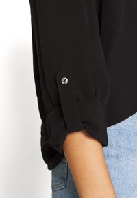 Svart blus med rullade ärmar och detaljer i silverknappar, tillverkad av ett mjukt tyg, kombinerad med ljusblå denim.