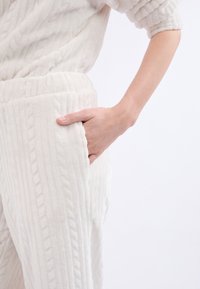 Conjunto de loungewear blanco y texturizado que presenta un diseño de punto de rombos. La mano de la modelo descansa en el bolsillo lateral de los suaves y cómodos pantalones.
