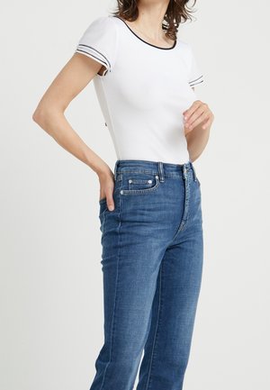 Femme portant un haut blanc à manches courtes avec des bordures noires et un jean bleu taille haute, posant avec une main dans la poche et l'autre près de la poitrine.