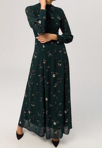Kvinna iklädd en lång mörkgrön blommig klänning med långa ärmar och svarta spetsiga skor, poserar med armarna korsade nära ansiktet.