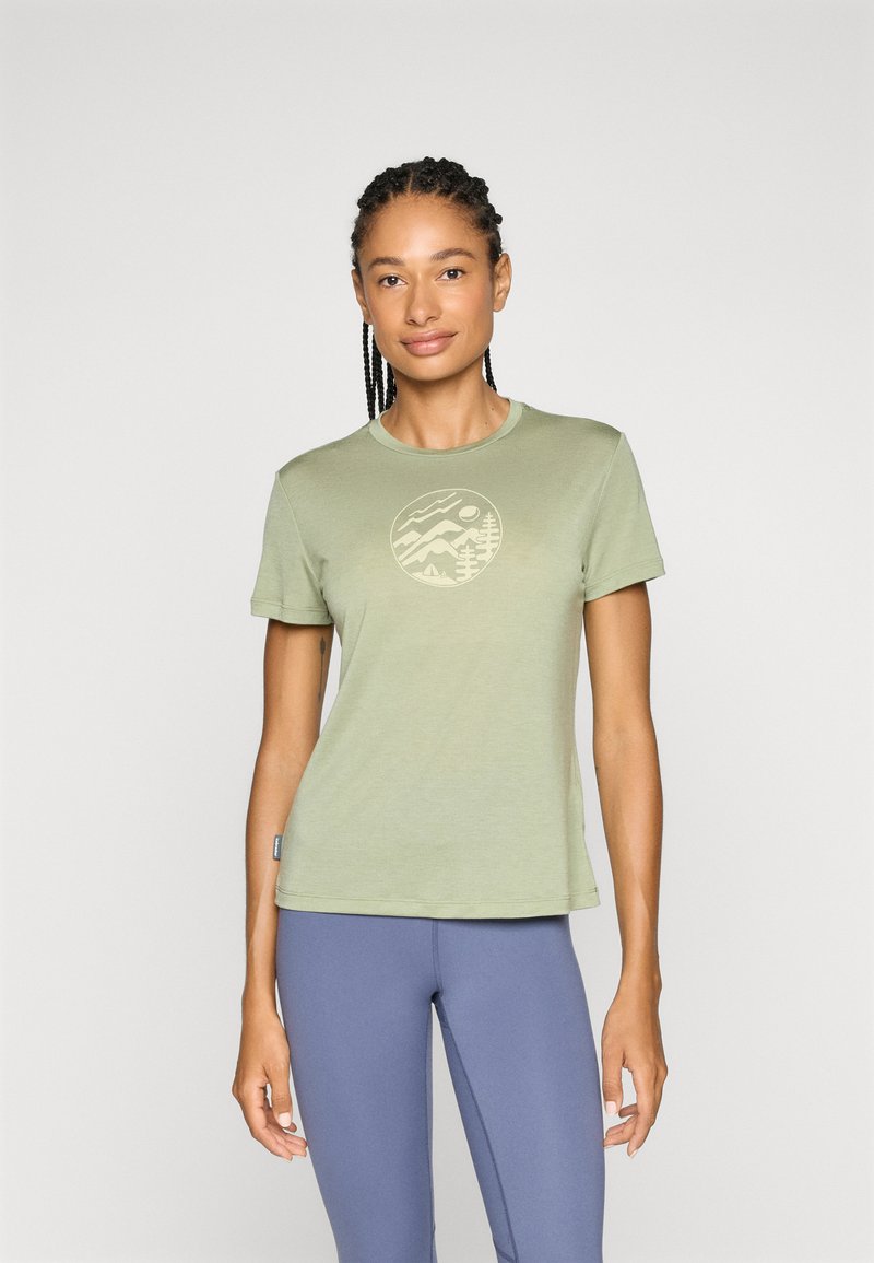 Frau mit geflochtenem Haar, die ein hellgrünes T-Shirt mit einem Grafikmotiv von Bergen und Bäumen sowie blaue Leggings trägt, steht vor einem schlichten Hintergrund.