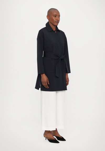 Ναυτικό παλτό με ζωνάκι, μακριά μανίκια, δύο μπροστινές τσέπες και γιακά, φορεμένο πάνω από φαρδιά λευκά παντελόνια και μαύρες γόβες.