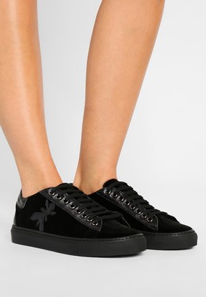 Czarne niskie sneakersy z sznurowanym designem i subtelnym logo mrówki z boku, noszone na bosych stopach na białym tle.