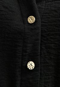 Tejido negro con un acabado texturizado, que cuenta con tres botones metálicos de superficie estampada en la parte delantera.