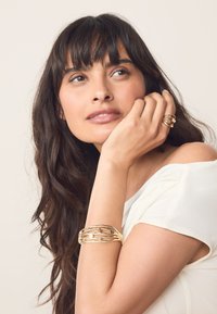 Gold-tone jewelry includes a multi-layered cuff bracelet with black bead accents and a matching ring. The model wears a white shirt.
