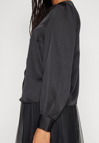 Haut en satin noir avec des manches bouffantes et un col arrondi, comportant une poche et une texture lisse. Associé à une jupe en tulle noire.