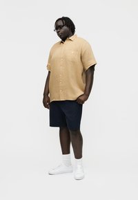 Prillidega mees, kes kannab beeži lühikeste varrukatega nööbitavat särk, tumesiniseid lühikesi pükse, valgeid sokke ja valgeid vabaajajalatsid, seisab ühevärvilise tausta ees.