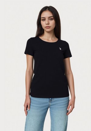 Mladá žena v čiernom krátkym tričkom s malým bielym logom na hrudi a svetlomodrých džínsoch stojaca pred jednofarebným pozadím.