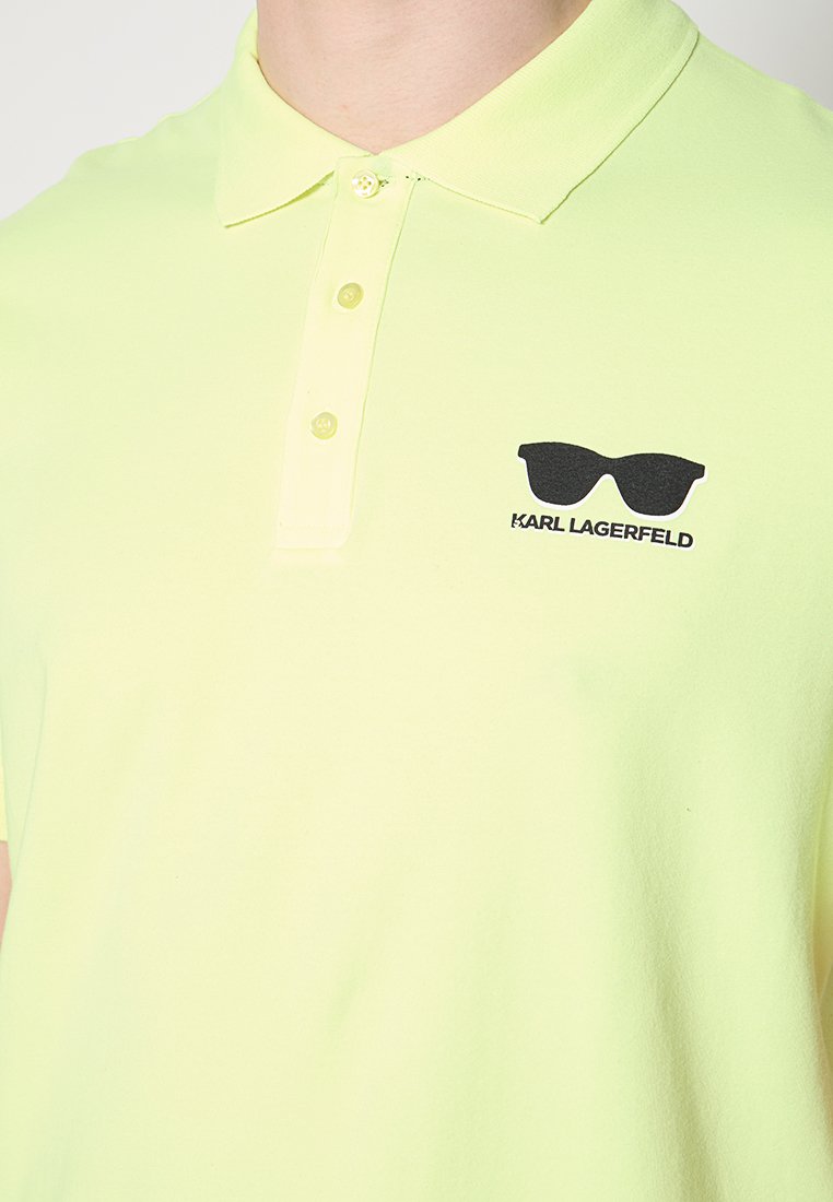 Mężczyzna w jasnobeżowej koszulce polo z logo czarnych okularów przeciwsłonecznych oraz napisem "Karl Lagerfeld" na piersi.