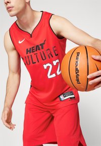 Camiseta de baloncesto roja con acentos negros, que presenta "HEAT CULTURE" en letras en negrita y texturizadas y el número "22." El jugador sostiene un balón de baloncesto naranja.