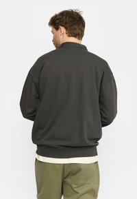 Donkergrijze sweatshirt met een relaxed fit, geribde manchetten en onderrand, en een eenvoudig ontwerp. G gedragen over een lichtgekleurde blouse en olijfgroene broek.