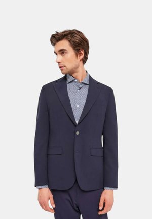 Marineblazer met een gesneden revers, enkele knoopsluiting en twee voorzakken. Daaronder is een gestreept shirt met contrasterende manchetten zichtbaar.