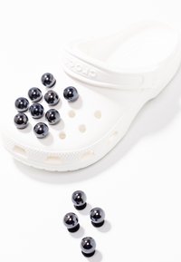 Des sabots Crocs blancs avec des trous de ventilation, ornés de plusieurs perles noires rondes sur le dessus et éparpillées à proximité sur un fond blanc.