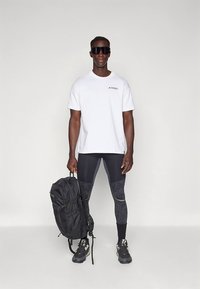 Hvit kortermet skjorte med "TERREX"-logo, svarte teksturerte leggings og en svart ryggsekk. Modellen har på seg mørke solbriller og tursko.