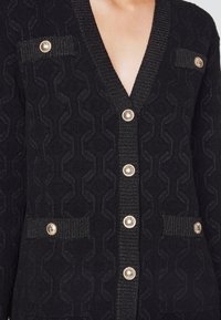 Cardigan noir texturé avec un éclat métallique, présentant des boutons dorés sur le devant et sur deux poches poitrine et deux poches hanches.