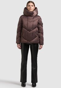 Braune Steppjacke mit hohem Kragen und Chevron-Muster, mit glänzendem Finish und Reißverschlusstaschen, kombiniert mit schwarzen ausgestellten Hosen.