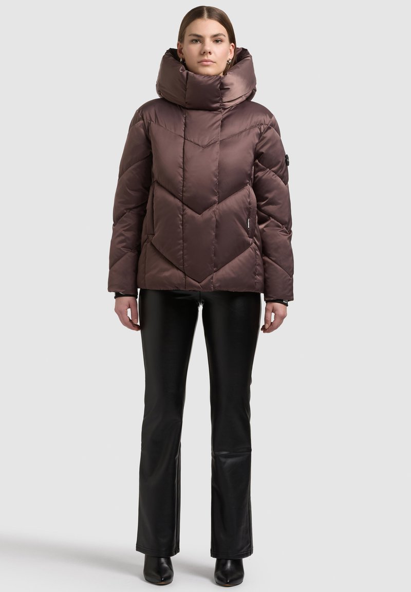 Braune Steppjacke mit hohem Kragen und Chevron-Muster, mit glänzendem Finish und Reißverschlusstaschen, kombiniert mit schwarzen ausgestellten Hosen.