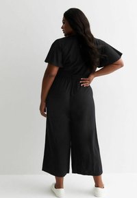 Svart jumpsuit med vida ben, korta ärmar och bälte i midjan. Tillverkad av lättviktigt tyg, med en nyckelhålsknäppning i ryggen. Vita sneakers.