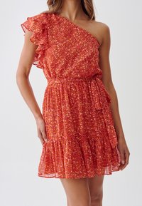 Robe florale rouge avec une épaule dénudée, manche froncée, taille ceinturée et ourlet froncé. Fabriquée en tissu léger et transparent.