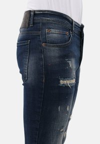 Donkerblauwe denim jeans met een slim fit, voorzien van versleten accenten, scheuren en een gestructureerde afwerking. Metalen klinknagels en een logopatch op de taille.