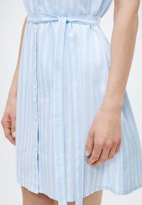 Φωτεινό γαλάζιο φόρεμα με κάθετες λευκές ρίγες, κουμπωτό μπροστινό μέρος και δεμένη ζώνη στη μέση, φορεμένο από πρόσωπο που απεικονίζεται από το μέσο του μηρού μέχρι τη μέση.