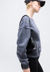 Νεαρός με γκρι φούτερ και μαύρο καπέλο που κρατά μικρή μαύρη δερμάτινη τσάντα ώμου, προφίλ πλευράς μπροστά σε λευκό φόντο.