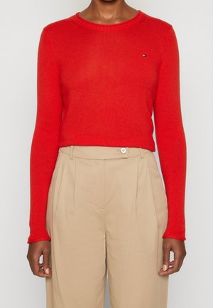 Personne portant un pull rouge ajusté à manches longues et un pantalon beige plissé taille haute, debout devant un fond uni.