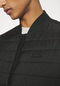 Giacca bomber nera trapuntata con una finitura testurizzata, polsini e colletto a costine. Presenta una patch logo discreta sul davanti. Chiusura con zip.