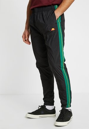 Mand iført sorte træningsbukser med grønne og gule sidestriber, sorte sneakers med hvide såler og en rød skjorte, stående med den ene hånd i lommen.