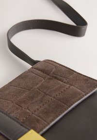 Sac bandoulière en cuir brun et daim avec un motif cousu texturé et une sangle fine réglable sur un fond clair.